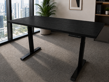 Load image into Gallery viewer, A black adjustable height standing desk with a wood-grain finish, positioned in a modern office setting with a large window, city view, and carpeted floor.