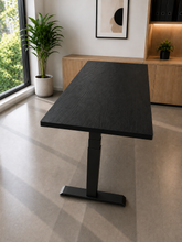 Load image into Gallery viewer, A modern adjustable height standing desk with a dark wood grain top and black metal pedestal base, shown in a bright, contemporary home office setting.