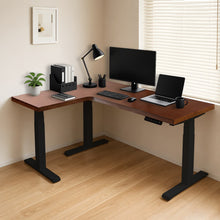 Load image into Gallery viewer, Walnut-finish L-shaped standing desk with black steel legs, shown with monitor, laptop and desk lamp