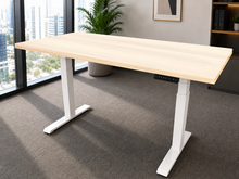 Load image into Gallery viewer, An adjustable height standing desk with a light natural wood top and white metal legs, shown in a modern office setting with a large window, gray carpet, and a potted plant.