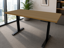 Load image into Gallery viewer, A modern adjustable standing desk with a light oak wood top and black metal legs, shown in a bright, contemporary office setting with a large window, city view, and carpeted floor.