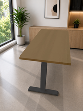 Load image into Gallery viewer, A modern adjustable height standing desk with a light brown wood-grain top and a dark gray metal T-style base, shown in a bright, contemporary home office setting.