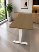 Load image into Gallery viewer, A modern adjustable height standing desk with a light brown wood-grain top and a white T-style metal base, shown in a bright, contemporary home office setting.