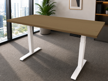 Load image into Gallery viewer, A modern standing desk with a light brown wood-grain top and white T-style legs, shown in a bright office setting with a window, city view, and potted plant.