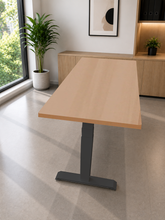 Load image into Gallery viewer, A modern adjustable height standing desk with a light wood grain top and a dark gray metal T-style base, shown in a bright, contemporary home office environment.
