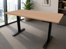 Load image into Gallery viewer, A modern adjustable height standing desk with a light natural wood top and black metal legs, shown in a bright office setting with a window, city view, and carpeted floor.